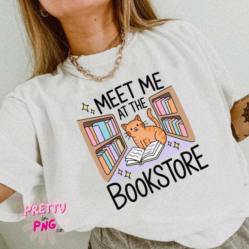 May include: White t-shirt with a graphic that says "MEET ME AT THE BOOKSTORE". The graphic features an orange cat reading a book in front of bookshelves with colorful books. The shirt has a relaxed fit.