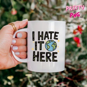 May include: White ceramic mug with the text "I HATE IT HERE" in black block letters. The "O" in "IT" is a blue and green globe. The mug is held by a hand with gold rings.