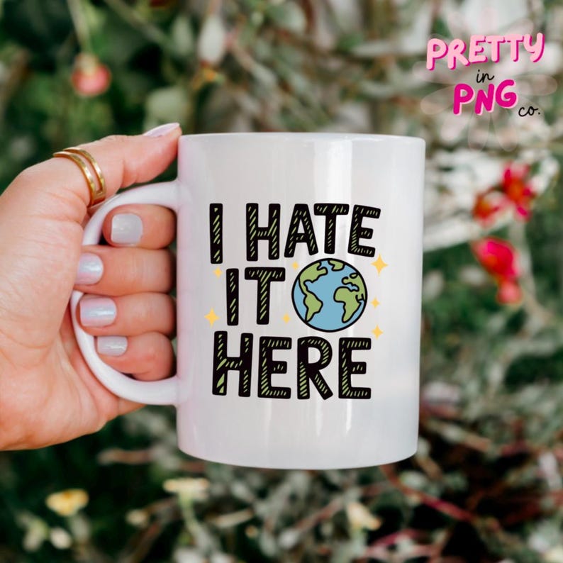 May include: White ceramic mug with the text "I HATE IT HERE" in black block letters. The "O" in "IT" is a blue and green globe. The mug is held by a hand with gold rings.