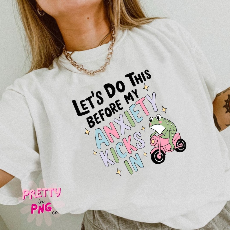 K&ouml;nnte beinhalten: Wei&szlig;es T-Shirt mit einem gr&uuml;nen Frosch, der ein rosa Motorrad f&auml;hrt. Der Text auf dem Shirt lautet "Let's do this before my anxiety kicks in".
