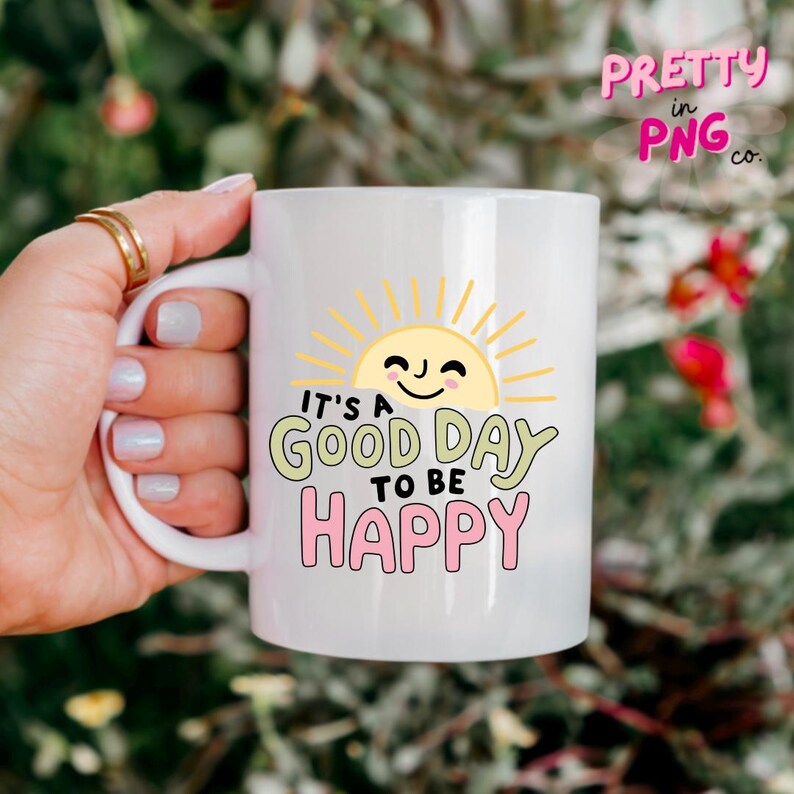 May include: White ceramic mug with a cheerful sun design and the text "It's a good day to be happy" in green and pink. The mug has a curved handle and is held by a hand with gold rings.