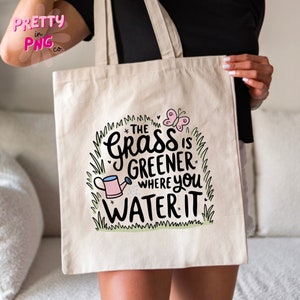 May include: A white canvas tote bag with a green grass and butterfly design. The text on the bag reads "The grass is greener where you water it."