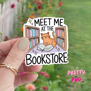 May include: A sticker with the text "MEET ME AT THE BOOKSTORE" featuring a cartoon orange tabby cat sitting on an open book in front of bookshelves. The sticker has a white border and is held by a hand.
