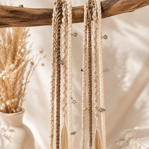 May include: Two sets of long, multi-strand hair extensions featuring a blend of cream, beige, and light brown braided synthetic fibers. The extensions are adorned with small metallic charms including suns, stars, and leaves, along with gold-toned beads.