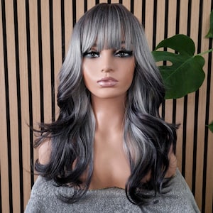 Puede incluir: Una cabeza de maniquí que muestra una peluca larga y ondulada con una mezcla de mechones grises, blancos y negros. La peluca presenta un flequillo recto y está peinada con rizos voluminosos. El maniquí está cubierto con una toalla gris, sobre un fondo de listones de madera.