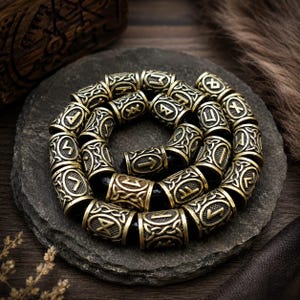 May include: A circular arrangement of cylindrical metal beads, each featuring an engraved runic character and intricate knotwork patterns. The beads are strung together on a dark cord, creating a continuous loop with a metallic gold finish.