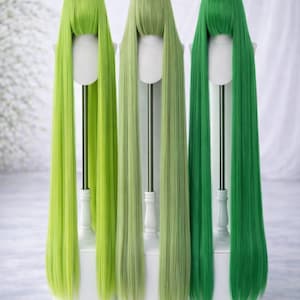 Puede incluir: Tres pelucas largas y lisas en tonos verdes se exhiben en soportes blancos. Las pelucas varían en color desde el verde lima claro hasta el verde bosque oscuro. Las pelucas están peinadas con flequillo y tienen la misma longitud.