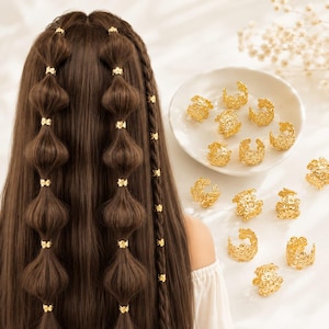 May include: A collection of small, gold-colored metal hair cuffs featuring intricate floral cut-out patterns. Several of these circular, open-ended hair accessories are displayed on a white plate, while others are worn on long brown hair.