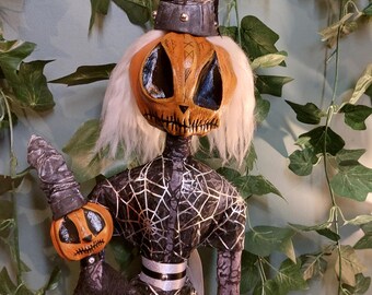 Może przedstawiać: Para handmade'owych lalek Halloweenowych z głowami dyni, jedna w czarnym kapeluszu czarownicy z srebrnymi akcentami, a druga trzymająca miotłę. Obie lalki są ubrane w czarną i srebrną tkaninę z wzorami pajęczyn.