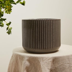 May include: A cylindrical ceramic planter featuring a dark charcoal gray finish and a vertical ribbed texture. The planter has a smooth, flat rim and a solid base, presenting a consistent, deep color throughout its exterior surface design.