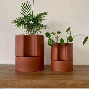 May include: Two terracotta-colored ceramic planters with a unique wavy design, each holding green plants. The planters are stacked on a wooden shelf, creating a modern and stylish look for indoor plants.