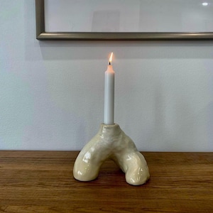 May include: A white ceramic candle holder shaped like a pair of legs with a single white candle burning in the centre.