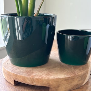 Puede incluir: Dos macetas de cerámica verde oscuro brillante de diferentes tamaños se encuentran sobre un soporte redondo de madera marrón claro. La maceta más grande contiene una planta verde con hojas grandes. Las macetas tienen un diseño moderno y minimalista.