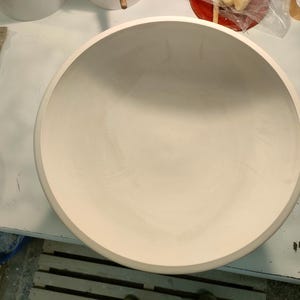 Peut inclure: Un grand bol en céramique blanc cassé de forme arrondie. Le bol a un large bord et une surface intérieure lisse. Le bol est posé sur une surface blanche, avec d'autres objets en céramique visibles en arrière-plan.