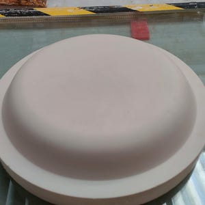 Peut inclure: Un objet rond en céramique rose clair avec un bord surélevé. L'objet a la forme d'un dôme et semble être un couvercle. L'objet est posé sur une surface en verre.