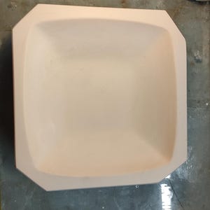 Könnte beinhalten: Eine weiße Keramikschüssel mit quadratischer Form und leicht abgerundeten Kanten. Die Schüssel hat eine glatte, glänzende Oberfläche und einen leicht konkaven Innenraum.