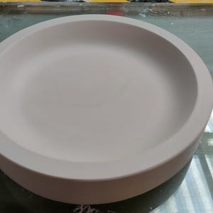 May include: A circular, off-white ceramic dish with a wide, shallow bowl and a thick, rounded rim. The dish has a smooth surface. The design is simple and modern, suitable for serving or display. The diameter is approximately 20 cm.