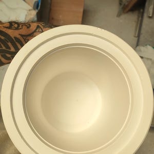 Könnte beinhalten: Eine cremefarbene Keramikschale mit breitem Rand und glattem Innenraum. Die Schale hat ein schlichtes, elegantes Design und scheint handgefertigt zu sein. Die Schale wird wahrscheinlich zum Servieren von Speisen oder als Dekorationsartikel verwendet.