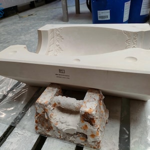 Könnte beinhalten: Weiße Keramikform mit einem detaillierten Blumendesign. Die Form ruht auf einem braunen, strukturierten Sockel. Die Form trägt den Text "WAGNER-WERKSTATT" und "OTTAWA, ONT. CAN."