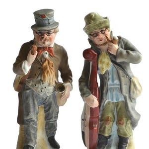 May include: Two porcelain figurines depicting musicians in vintage attire. The left figure wears a grey top hat, brown jacket, and holds a small instrument. The right figure wears a green hat, grey coat, and holds a large red string instrument.