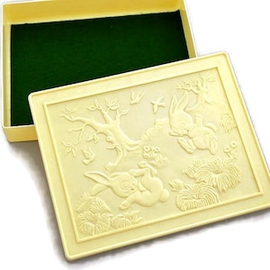 May include: A light yellow rectangular box with a lid featuring a bas-relief design of rabbits, trees, and flowers. The box interior is lined with dark green fabric. The lid is partially open, revealing the interior.