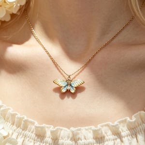 Puede incluir: Un collar de tono dorado con un colgante de mariposa con alas de esmalte azul claro y blanco y un pequeño acento de perla central. La delicada cadena descansa sobre una prenda de tela color crema, resaltando los detalles de la mariposa.