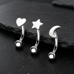 Può includere: Tre piercing per sopracciglia argentati con forme diverse: un cuore, una stella e una luna crescente. Ogni anello ha una barra curva con una piccola sfera a ciascuna estremità. Le forme sono lisce e lucide.