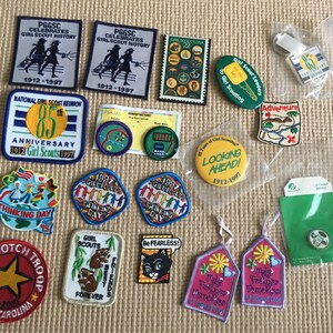 May include: A selection of Girl Guide memorabilia, including badges, pins, and tags. Badges display text such as "Girl Scout History," "Thinking Day," and "Top Notch Troop." Colours include blue, green, red, and yellow. Some items are in plastic packaging.