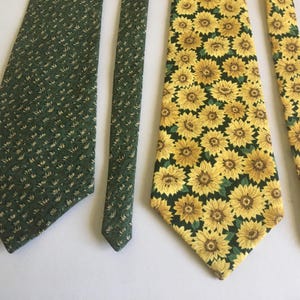 May include: Two neckties. One is dark green with a small, repeating pattern. The other is yellow with a pattern of sunflowers. Both ties are made of fabric and are displayed on a white surface.