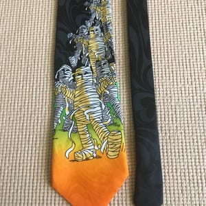 May include: A Halloween-themed necktie featuring a design of mummies walking across a gradient of yellow and orange. The top portion of the tie is black with a stylized orange moon and decorative swirls.