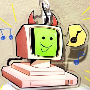 May include: A clear acrylic charm featuring a cartoon computer with a smiling face on the green screen and red devil horns. A musical note is on a yellow speech bubble, with blue musical notes and yellow lines radiating from the charm.