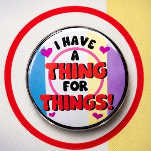 May include: A round pinback button featuring a vertical striped background in light blue, white, yellow, and purple. The text "I HAVE A THING FOR THINGS!" is printed in black and red, surrounded by four small pink hearts and a thin pink circle.