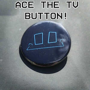 May include: A dark blue button with the text "ACE THE TV BUTTON!" in white, pixelated font. The button features a blue graphic of two squares and a line. The button is round and has a glossy finish.
