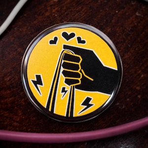 Puede incluir: Un botón redondo con fondo amarillo que presenta una mano negra agarrando tres líneas amarillas. Encima de la mano hay tres corazones negros y rayos a los lados. El botón tiene un acabado transparente y brillante.