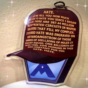 Może przedstawiać: Przezroczysty akrylowy brelok do kluczy z głową robota w stylu baseballowej czapki. Czapka jest ciemnobrązowa ze słowem "HATE" i długim cytatem wydrukowanym w kolorze złotym. Twarz robota jest niebieska z białym emblematem.