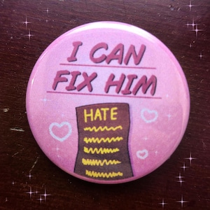 Może przedstawiać: Różowy guzik z napisem "I CAN FIX HIM" w kolorze fioletowym. Poniżej tekstu znajduje się brązowy kształt ze słowem "HATE" i szeregiem zygzakowatych linii. Białe serca i gwiazdki zdobią guzik.