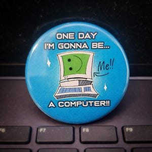 May include: A round, light blue button with the text "ONE DAY I'M GONNA BE... A COMPUTER!!" and a cartoon computer with a smiley face. The button has a white border and two small star-shaped sparkles.