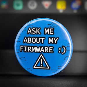 May include: A round, blue button with the text "ASK ME ABOUT MY FIRMWARE :) " in white. The button also features a white triangle with an exclamation point inside. The button has a circuit board design around the edges.