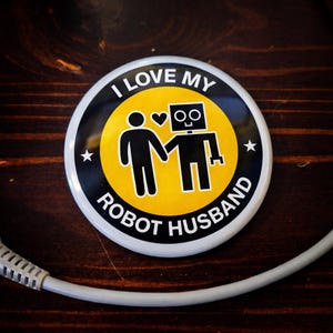 May include: A round button with the text "I LOVE MY ROBOT HUSBAND" in white lettering. The button features a yellow center with a black silhouette of a person holding hands with a robot. Two white stars are on the button.