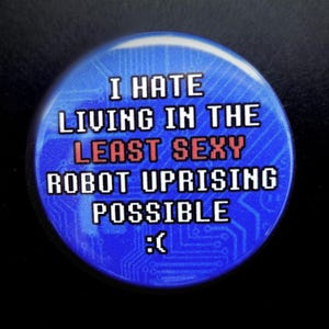 Puede incluir: Una chapa redonda con fondo azul y un patrón de placa de circuito. El texto dice: I HATE LIVING IN THE LEAST SEXY ROBOT UPRISING POSSIBLE seguido de un emoticono triste. La palabra SEXY está en rojo, mientras que el resto del texto es blanco.