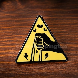 Könnte beinhalten: Eine dreieckige Emaille-Anstecknadel mit gelbem Hintergrund und schwarzem Rand. Das Design zeigt die schwarze Silhouette einer Hand, die ein Kabel greift, umgeben von drei kleinen schwarzen Herzen oben und zwei schwarzen Blitzen unten.