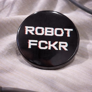 May include: A black round button with the text "ROBOT FCKR" in white, with a glitch effect. The button is on a light-colored fabric, with a gray cable in the background.