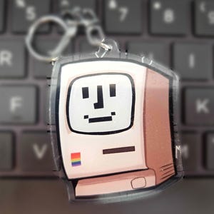 Puede incluir: Un llavero de acrílico transparente con la ilustración de un monitor de computadora beige retro, un rostro pixelado en la pantalla y un pequeño icono cuadrado de arcoíris en la esquina. Incluye una anilla de metal en la parte superior.