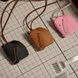 May include: Three leather elephant keyrings in black, brown, and pink. Each keyring has a leather strap and is displayed on a grid-lined surface. A metal tool and spools of thread are also visible.
