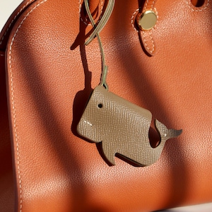 Könnte beinhalten: Eine braune Ledertasche mit einem hellbraunen, walförmigen Lederanhänger, der am Henkel befestigt ist.