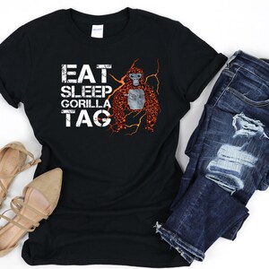 May include: Black t-shirt with the words "EAT SLEEP GORILLA TAG" in white block letters. A graphic of an orangutan with orange and brown fur is on the right side. The shirt is paired with blue denim jeans and tan sandals.
