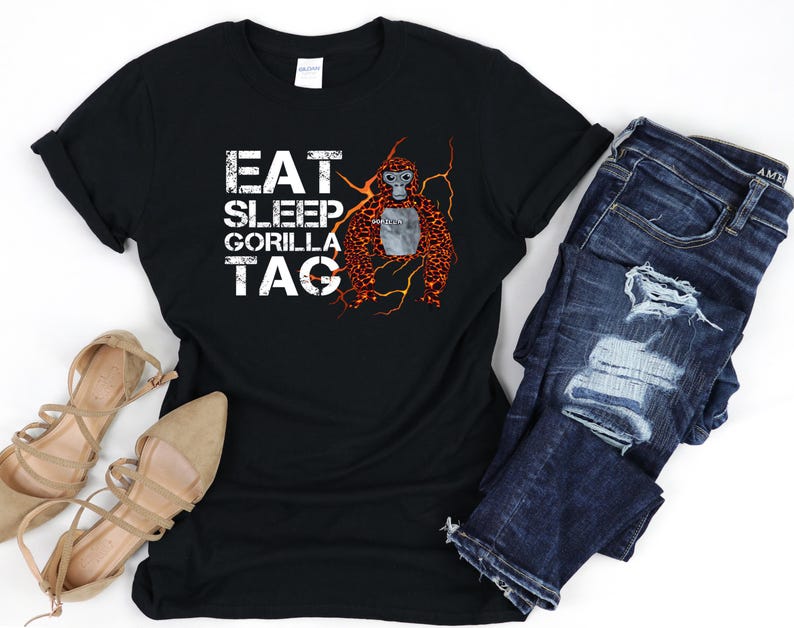 May include: Black t-shirt with the words "EAT SLEEP GORILLA TAG" in white block letters. A graphic of an orangutan with orange and brown fur is on the right side. The shirt is paired with blue denim jeans and tan sandals.
