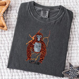 May include: Dark gray Comfort Colors t-shirt featuring a cartoon gorilla design with orange and black fur, a gray face, and the word "GORILLA" on its chest. The phrase "LET'S GO" is printed below the gorilla.