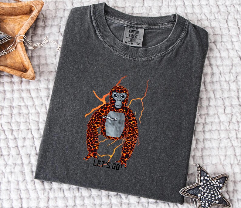May include: Dark gray Comfort Colors t-shirt featuring a cartoon gorilla design with orange and black fur, a gray face, and the word "GORILLA" on its chest. The phrase "LET'S GO" is printed below the gorilla.