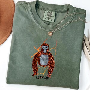 May include: An olive green t-shirt featuring a graphic of a gorilla with orange and brown fur and the text "LET'S GO". The shirt also has the text "GORILLA" on the chest. A gold necklace with a pendant is draped across the shirt.